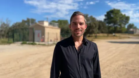 A smiling man with a beard and moustache wearing a black shirt stands on a sunny road in the south of France