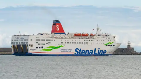 A white Stena Line ferry leaves port. The ship has a red and blue chimney with a white 'S'. 'Stena Line' is written across the side of the ship in blue.