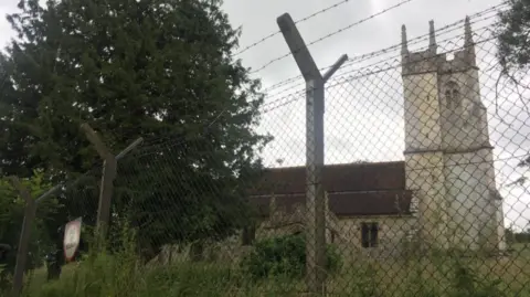 A church with a tower stands behind a tall mesh wire fence with rows of barbed wire at the top