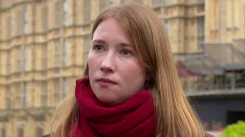Helena Dollimore stă în fața Palatului Westminster. Poartă o haină închisă la culoare și o eșarfă roșie.