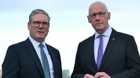 Sir Keir Starmer, with grey hair and  glasses, stands alongside John Swinney, bald with glasses, with an Edinburgh skyline in the background. Both are wearing dark suits and white shirts. Starmer is wearing a green tie while Swinney is wearing a purple one. 