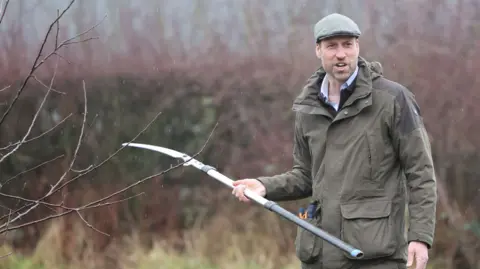 The Prince of Wales, dressed in a green jacket, open-necked shirt, navy trousers and flat cap, holds a scythe outside next to a tree. He is grinning at someone off camera.