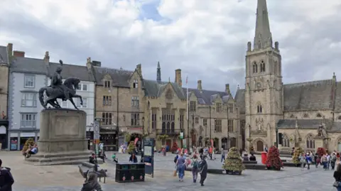 Google Durham Market Place, where the Town Hall is located, alongside St Nicholas Church and the statue of Charles William Vane on horseback. It is a paved area with a few dozen people milling about. The buildings are old, medieval in style.