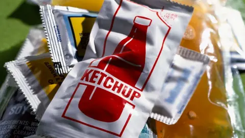 A tomato ketchup sachet, with a red bottle on a white background, lies on top of various other sachets of condiments