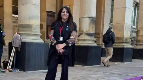 Shabina Bano outside Birmingham City Council House. She is wearing black trousers and a black shirt and smiling at the camera. She has long black hair.