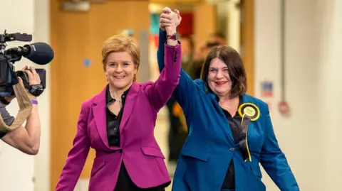 PA Media Nicola Sturgeon and Susan Aitken hold hands lifted into the air in a show of triumph. Sturgeon has short fair hair and wears a pink blazer with a black shirt, and Aikten has shoulder-length dark brown hair and wears a blue blazer, black shirt, and yellow and black SNP rosette. A TV camera films them