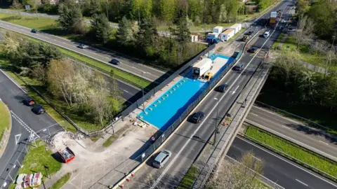 An aerial view of a bridge as it crosses a dual carriageway. One side of the carriageway is closed and is covered in blue tarpaulin with the slip road onto the dual carriageway below also closed. 