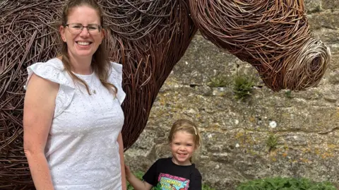 BBC A woman wearing a white top, with a small girl stood next to her. They are stood in front of a wicker sculpture of a horse.