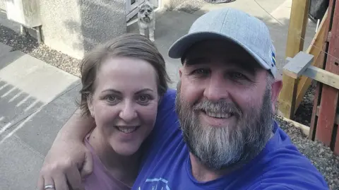 A man with a longer beard is standing in front of a house with his arm around a woman with short blond hair. She is wearing a pink top, while he is dressed in a dark-blue t-shirt and a grey cap. There is a schnauzer dog visible behind the couple.