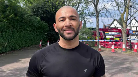 Kate Bradbrook/BBC A man with a beard and a black t-shirt is stood in front of Pink Punters which has red barriers, cone and a blue police tape restricting access