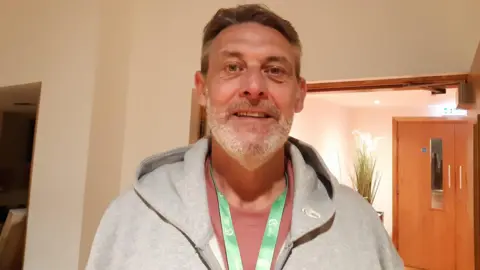 Malcolm Roberts Malcolm Roberts wearing a grey hoodie and green lanyard