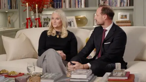 NRK A woman with a black top and blonde hair sits on a sofa, as a man to her right with a beard looks on