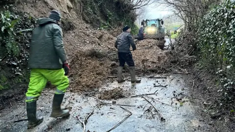 Two men in protective clothing and wellington boots clean a road covered in mud.