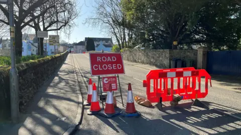 A 'road closed' sign stands surrounded by traffic cones, alongside a pair of red barriers which are blocking the approach to Gote Bridge in Cockermouth. Trees line the approach to the stone bridge, with a broad blue gate to the right and a town in the distance.