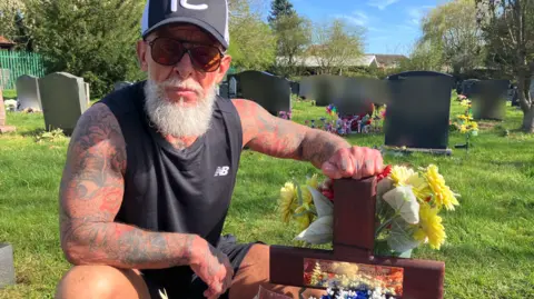 A person wearing a cap and sunglasses kneels beside a flower-covered grave marker in a cemetery.
