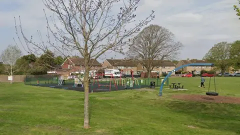 A small play area in a green field with eight houses in the background. There is a small brown tree in the foreground