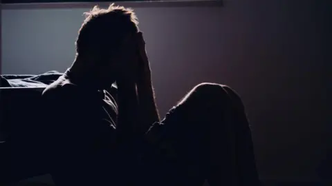 silhouette stock image of a man sat on the floor with his head in his hands, appearing in distress