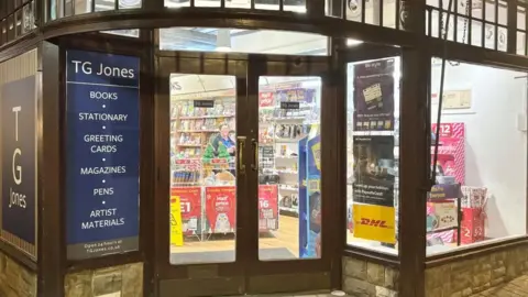 The front of a shop showing wooden-framed glass doors, a wooden-frame glass window and a blue rectangular sign, headed TG Jones and advertising: "BOOKS, STAIONARY, GREETING CARDS, MAGAZINES, PENS, ARTIST MATERIALS".