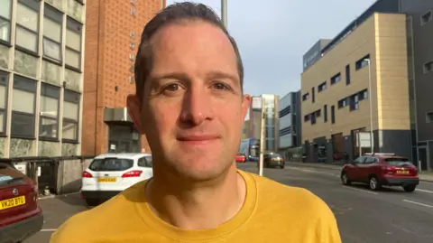 Tom Percival/BBC Rob Pardon smiles and looks directly at the camera wearing a yellow T-shirt. He is photographed outside on a pavement, with a street behind him. 