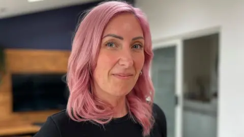 A view of a woman with pink shoulder length hair in her home, looking at the camera for a photo.