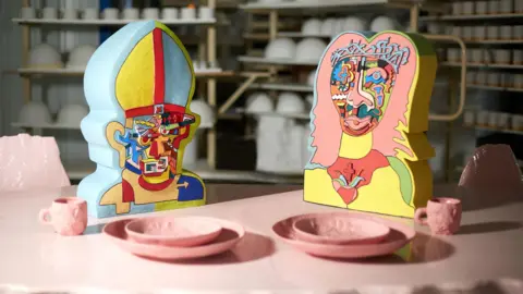 Two multi-coloured clay busts of the heads of Jesus and The Pope sitting on a pink table with a pink tea set in front.