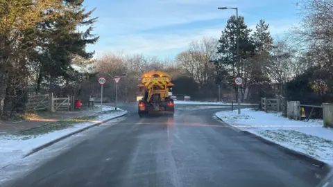 Shaun Whitmore/BBC The back of a yellow gritter lorry, with a yellow and red striped sign that says "Spreading". It is at a T-junction, with the verges mostly covered in snow.
