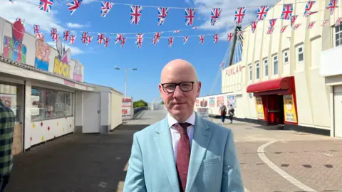  Patrick Hurley wears a blue suit and claret tie - he has dark rimmed glasses and a bald head. Union flags bunting is above his head at the entrance to the pier which is boarded up further down. 