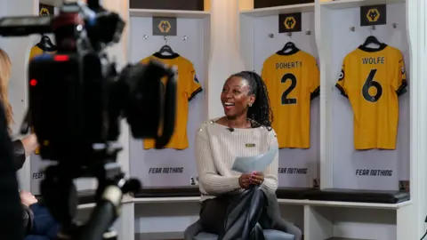 Wolves Foundation A woman with her hair in a ponytail, wearing a beige jumper and black leather trousers is holding a white piece of paper as she smiles to the left. A TV camera is pointed in her direction 
