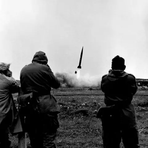 Getty Images Three men look through binoculars from behind a wire fence as a missile takes off. There are flames emerging from the rocket and a plume of smoke.