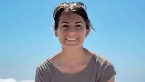 Family handout image of Lisa Ross - Close-up portrait of a person outdoors against a clear blue sky, wearing a grey short-sleeved top with sunglasses resting on the top of the head. The image is brightly lit by sunlight, with a small section of white cloud visible in the lower left corner.