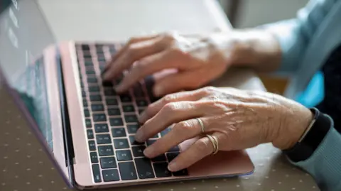 An elderly woman is typing on a laptop. She is wearing rings. She's wearing a blue cardigan and a digital watch. The table has a grey spotty cover. 