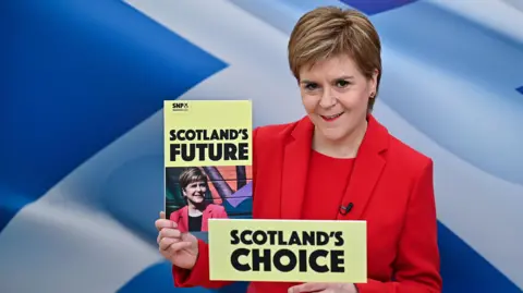 PA Media Nicola Sturgeon - a woman with short brown hair, wearing an all red outfit - holds up a booklet saying Scotland's Future while standing behind a sign saying Scotland's Choice