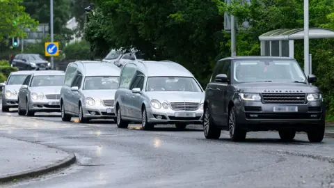 A convoy of silver hearses and mercedes, led by a black Range Rover driving along a road