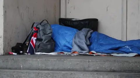 An unidentified homeless person sleeping rough in a blue sleeping bag in a doorway. A grey rucksack is in front of the person.