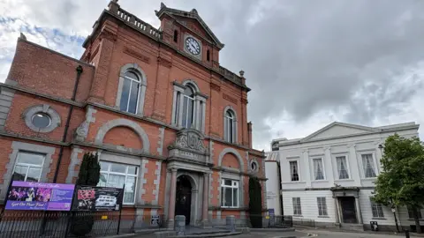 A side-on view of Newry Town Hall with the neighbouring Sean Hollywood Arts Centre at a perpendicular angle, facing the camera. The town hall is a red-bricked, two-story building with a clock on the very top section, large symmetrical windows and a two decorative port-hole windows at either side. The smaller arts centre has a grey-cream neoclassical facade.