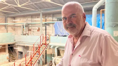 A man with a trimmed beard and moustache, wearing an open necked pink shirt. He is smiling at the camera, and behind him is a large two storey industrial space with stairs and pipework on display