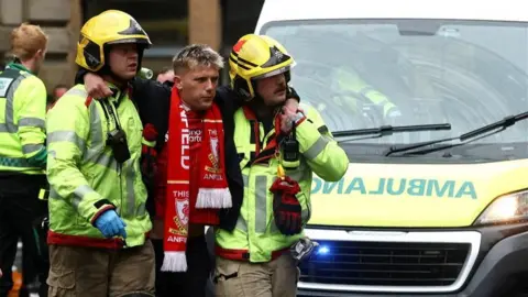 Reuters A man wearing a Liverpool football scarf is supported has his arms wrapped around two paramedics who are helping him to walk