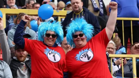 Two women are dressed in costumes similar to Thing One and Thing Two characters from Dr Seuss's Cat in the Hat books. They wear blue wigs with red tops and sunglasses One of the women's tops says Amy 1 and the other says Amy 2. They are standing in football stadium stands with their arms raised in celebration as they smile at the camera.