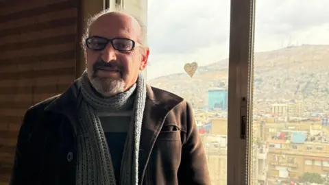 Lawyer Hussein Issa, standing by the window in a jacket and scarf, looking back at Damascus - he believes many Assad-era judges should be expelled from the courts.