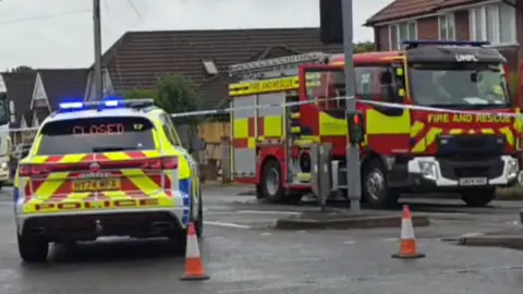 A police car sat in the road, parked in front of a police cordon with officers stood on the pavement. There is an ambulance with a lorry behind it. Also there is a fire engine on the right of the image.