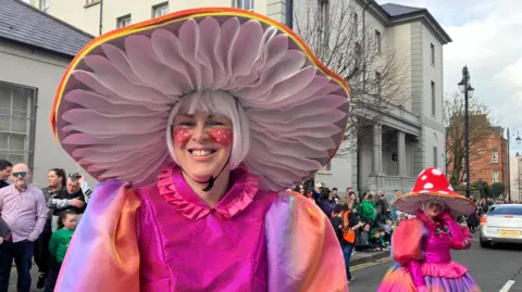 Two people in living costumes on the subject of mushroom take part in a parade. The first wears a large pink mushroom hat and a pink dress with dazzling sleeves, while the second person attracts a red mushroom hat with white spots and a multicolored dress in the background. The street is lined with spectators and contributes to the festive atmosphere.