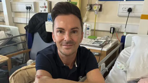 Northampton General Hospital A man in a navy polo leans over a hospital bed. He has dark hair with a large quiff.