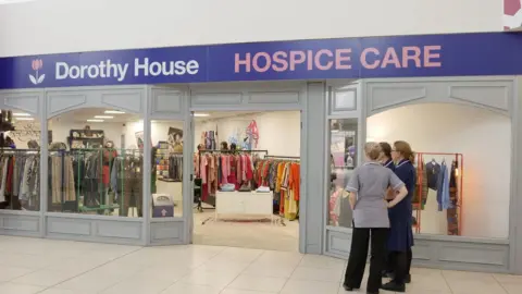 Dorothy House Hospice Care The frontage of a Dorothy House shop inside a shopping centre. Three women in nurses uniform stands chatting outside. There are lots of colourful garments on rails to be seen through the large glass panes.