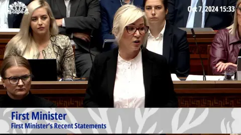 Northern Ireland Assembly Michelle O'Neill - a woman with blonde hair in an updo wears red-framed glasses. She is standing with other MLAs around her and she is wearing a black suit jacket and white buttoned shirt.