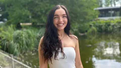 A woman in a cream strapless top with long black hair smiles broadly as she stands in front of a lake in a park. A glass sided building can be seen behind her.