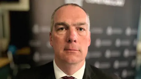 Martin Giles/BBC Det Supt Phill Gray slightly smiles at the camera. He has dark and grey hair that has been shaved closely to his head. He wears a black suit with a white shirt and red tie. The background behind him is blurred.