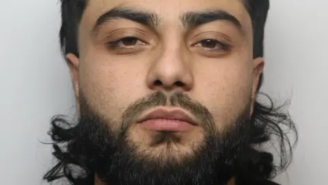 West Yorkshire Police Mugshot of Luqman. He is an Asian male and has dark hair and facial hair. Stood up against a grey wall.
