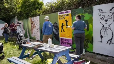 The Green Backyard People standing and painting artwork on a wall. The two nearest the camera have their back turned. There is a bench in the foreground.