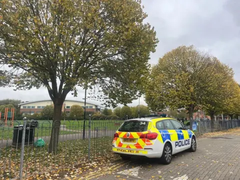 Bede Park - a leaf-strewn pathway, with a police car parked by railings and a grassed playing field