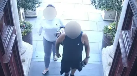The Custom House A woman in a white sweatshirt and leggings and a man in a black vest and black shorts walking into a building with red doors. Both of their faces are blurred.